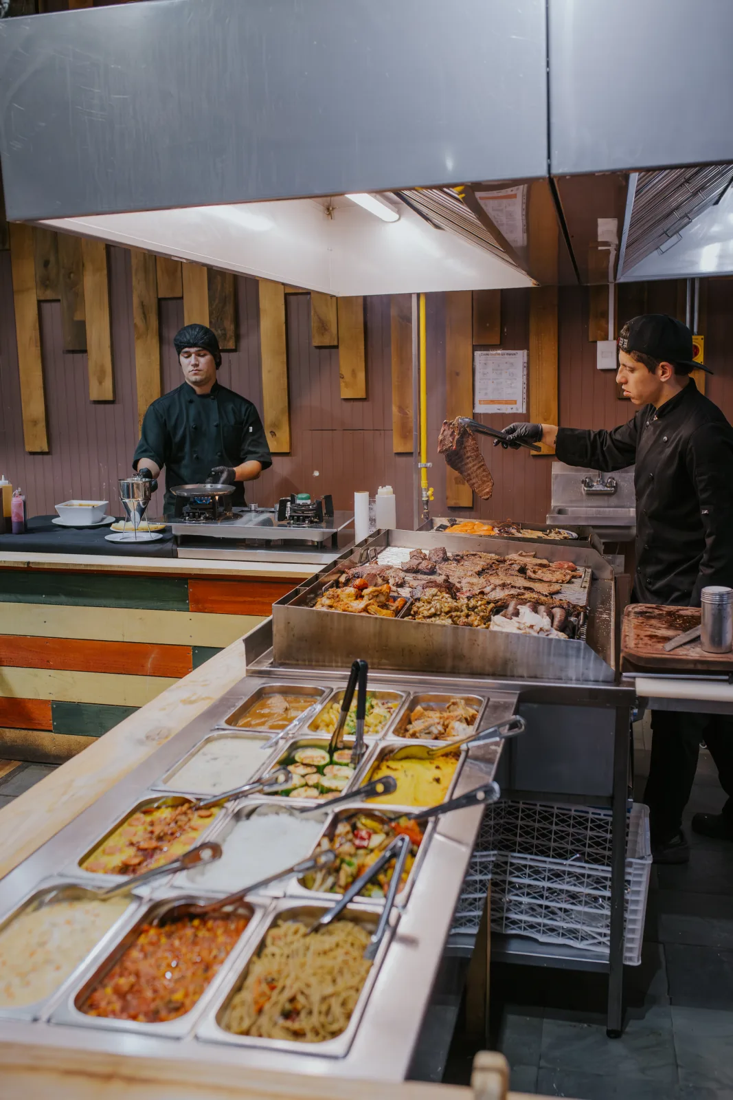Panorámica de la estación de parrilla y barra de guarniciones calientes atendida por dos cocineros del restaurante