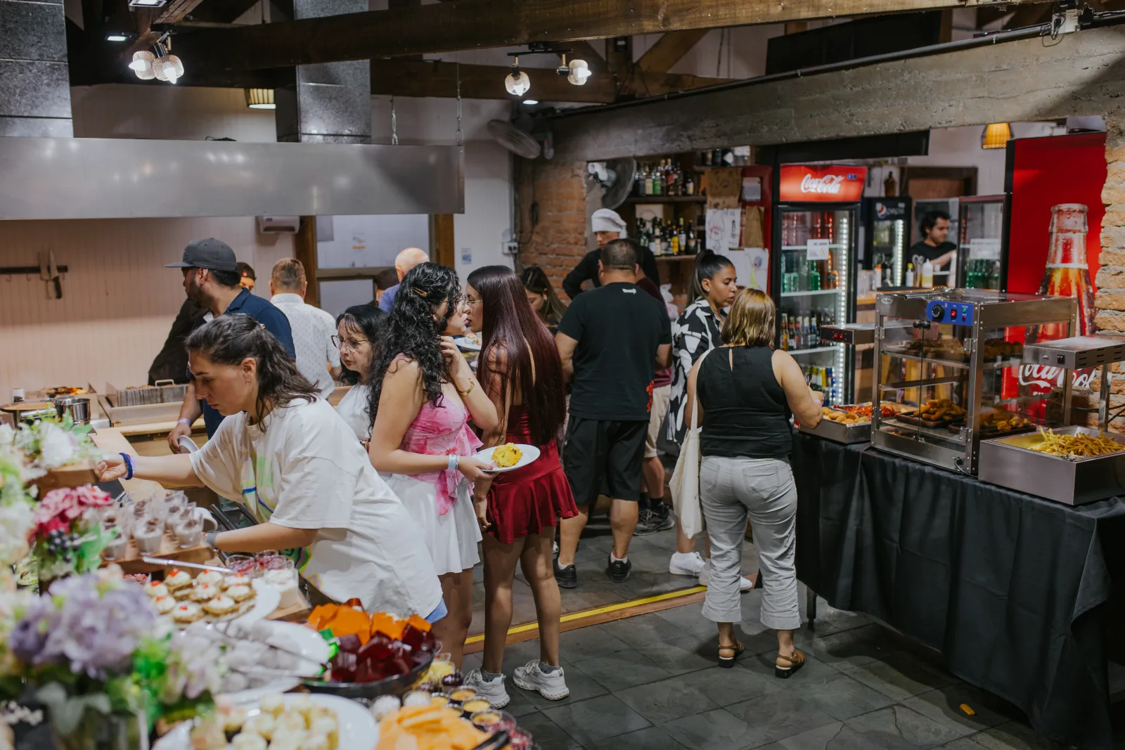 Personas recorriendo el buffet con platos junto a refrigerador de bebidas en ambiente cálido