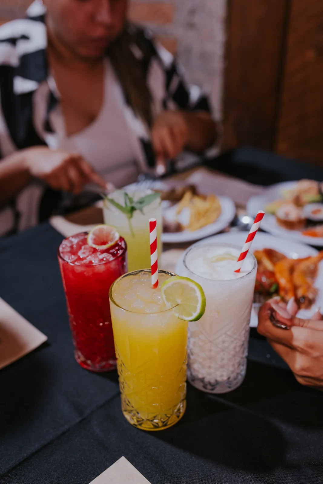 Tres bebidas refrescantes de colores con bombillas y limón sobre mesa con platos de fondo