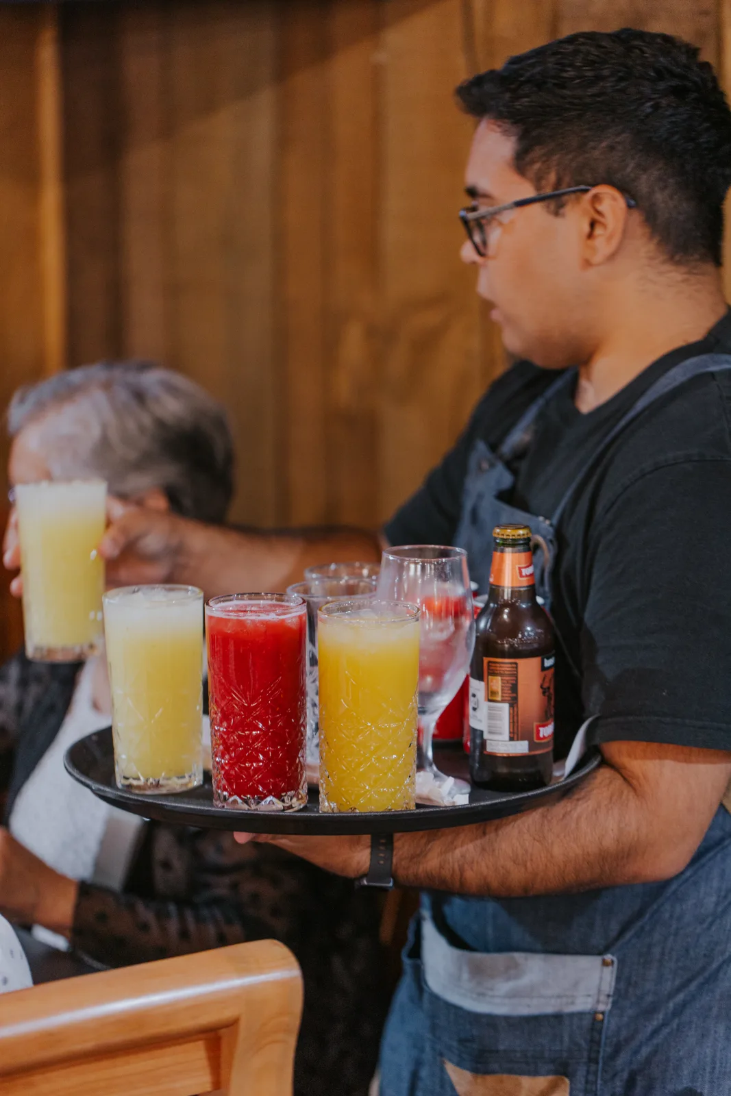 Mesero con delantal lleva bandeja de jugos naturales y cerveza