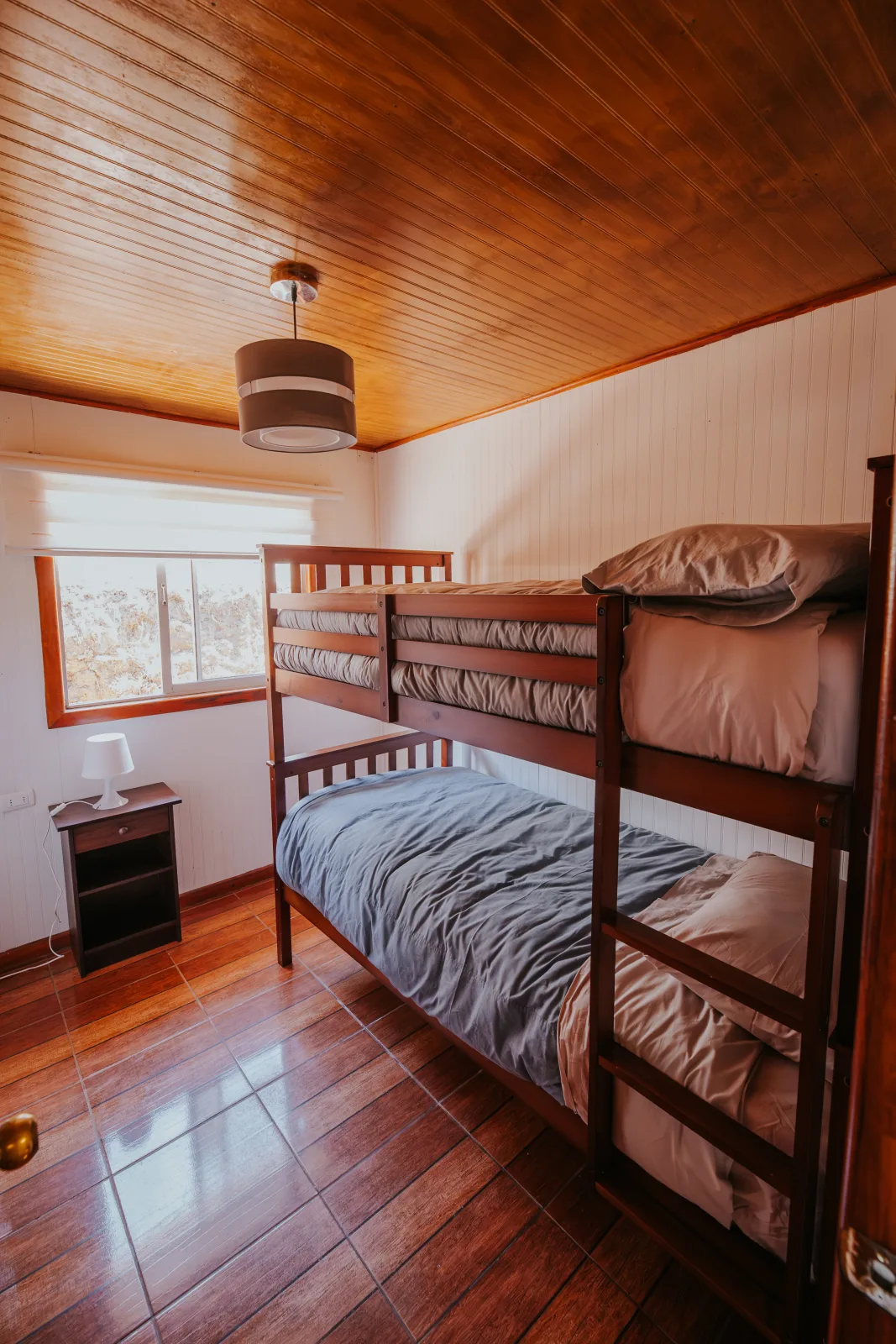 Habitación con litera de madera, ropa de cama gris y velador junto a la ventana