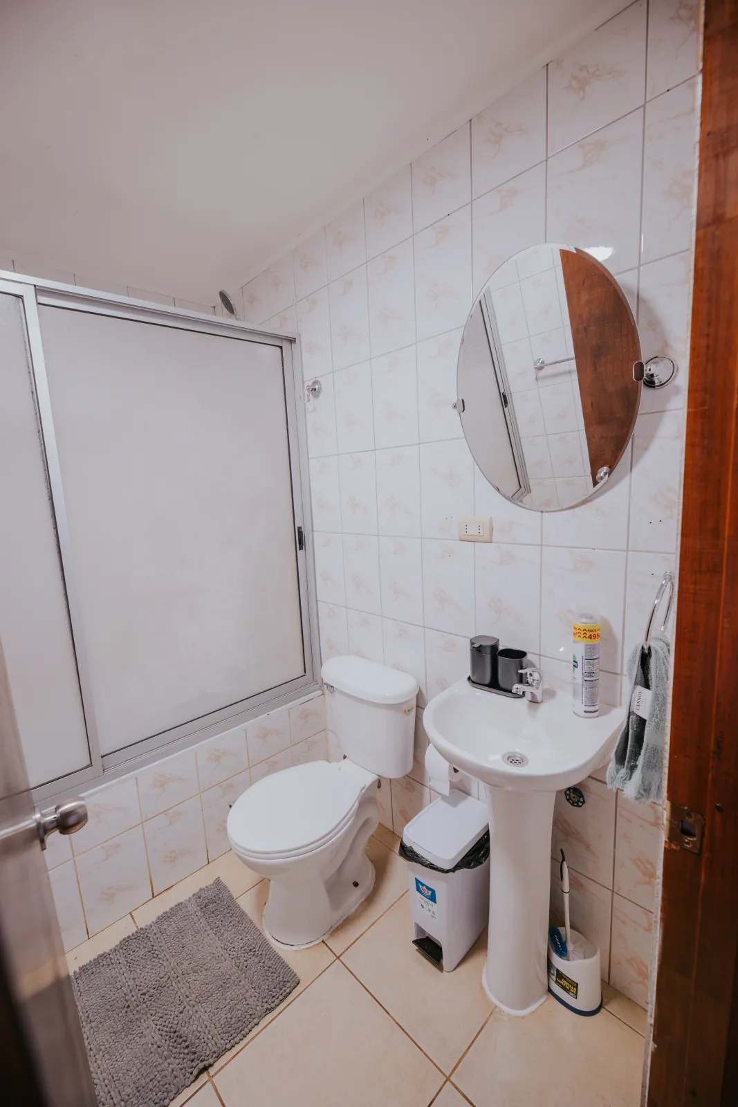 Baño limpio con ducha de vidrio, inodoro, lavamanos y espejo redondo