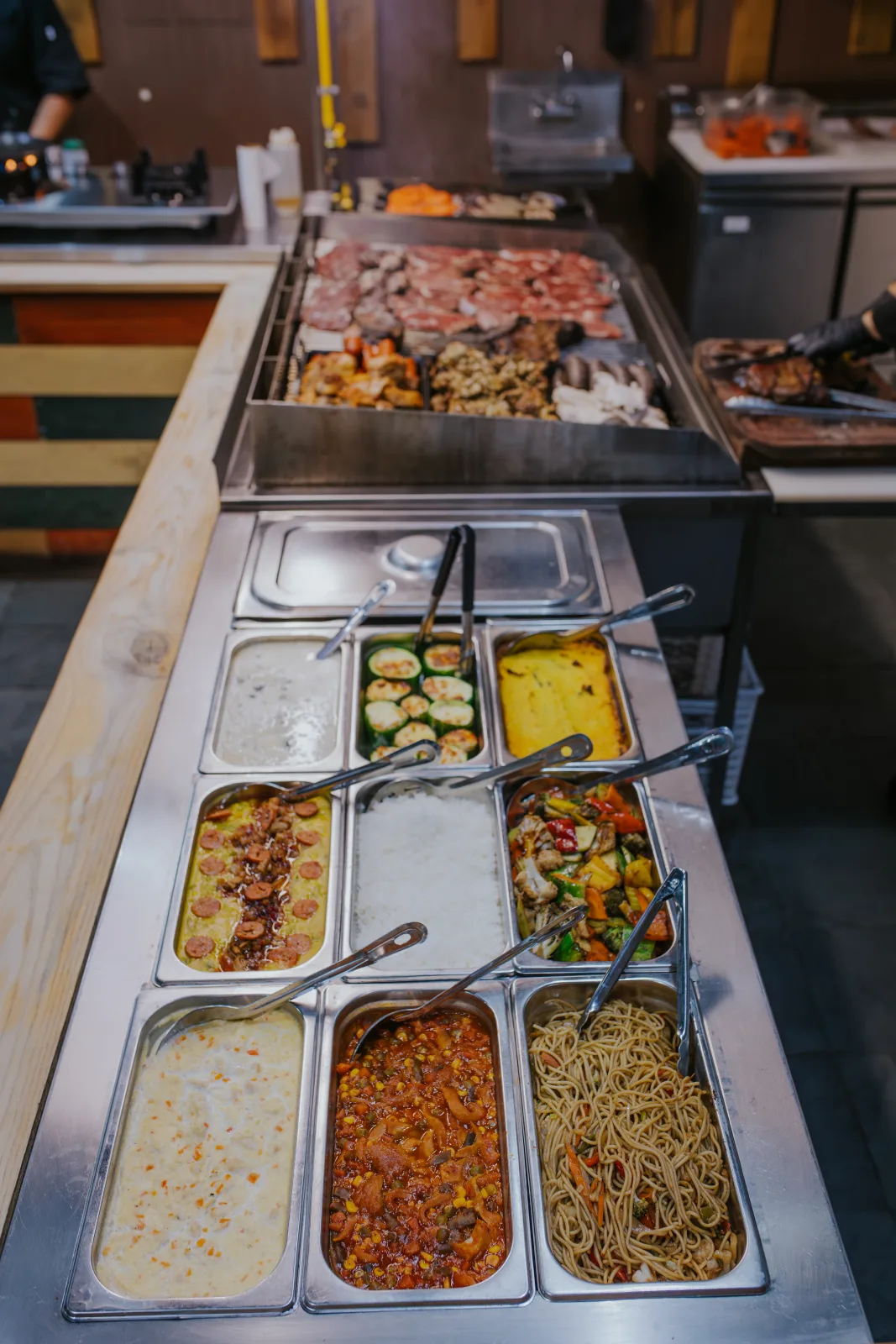Barra buffet de guarniciones calientes con bandejas de zapallitos, fideos y guisos, parrilla de carnes al fondo