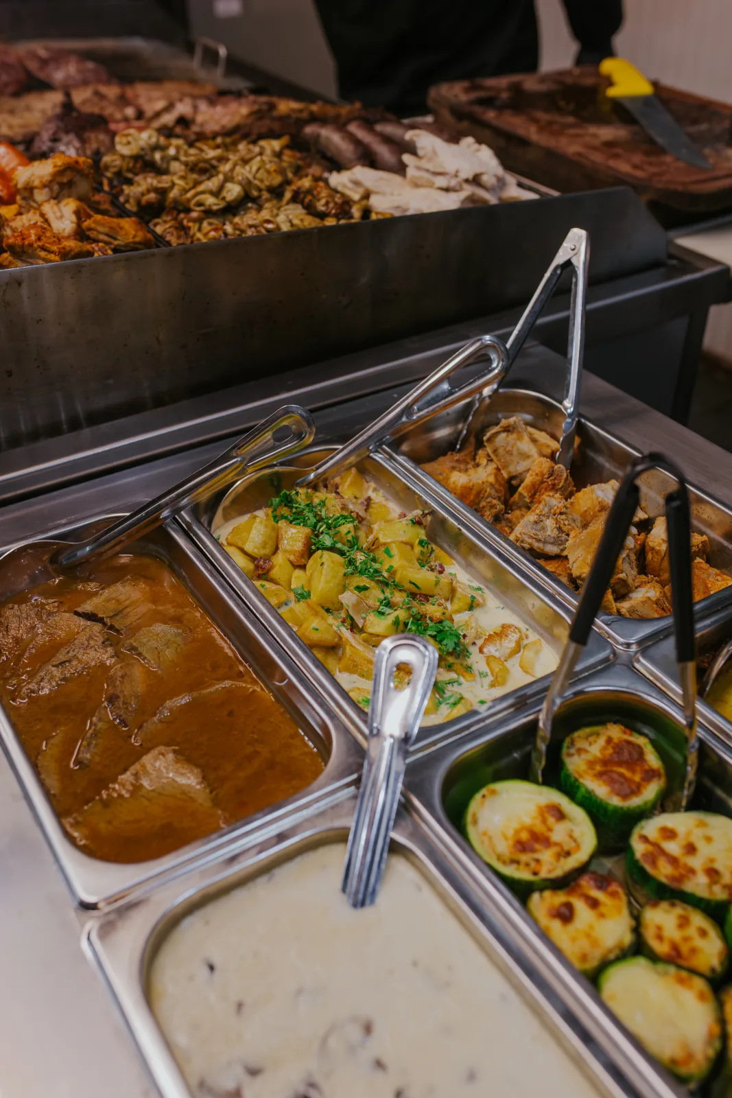 Bandejas de buffet con salsas, zapallitos grillados, papas y carnes en la estación de guarniciones calientes
