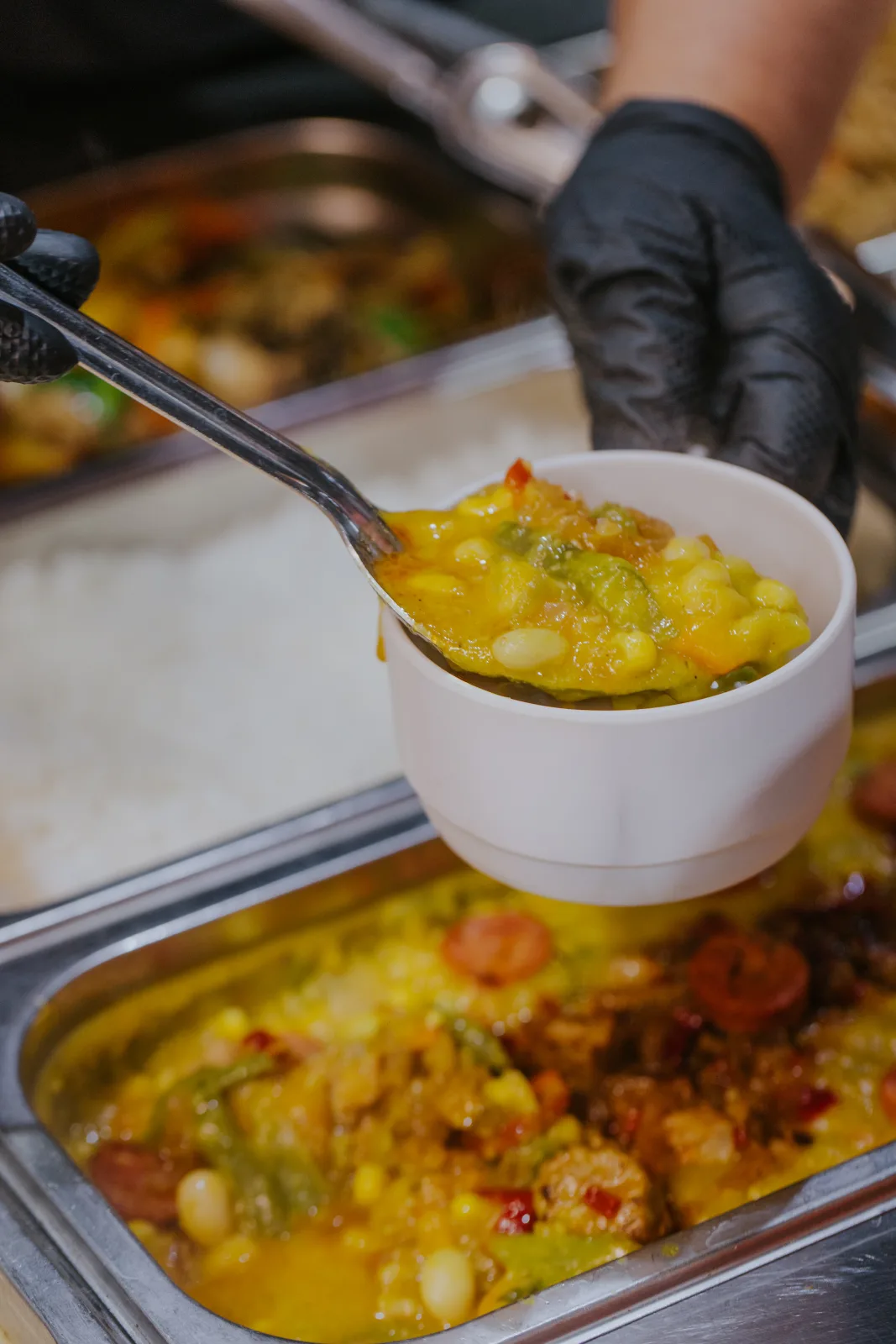 Mano enguantada sirviendo guiso caliente de porotos con chorizo y choclo del buffet
