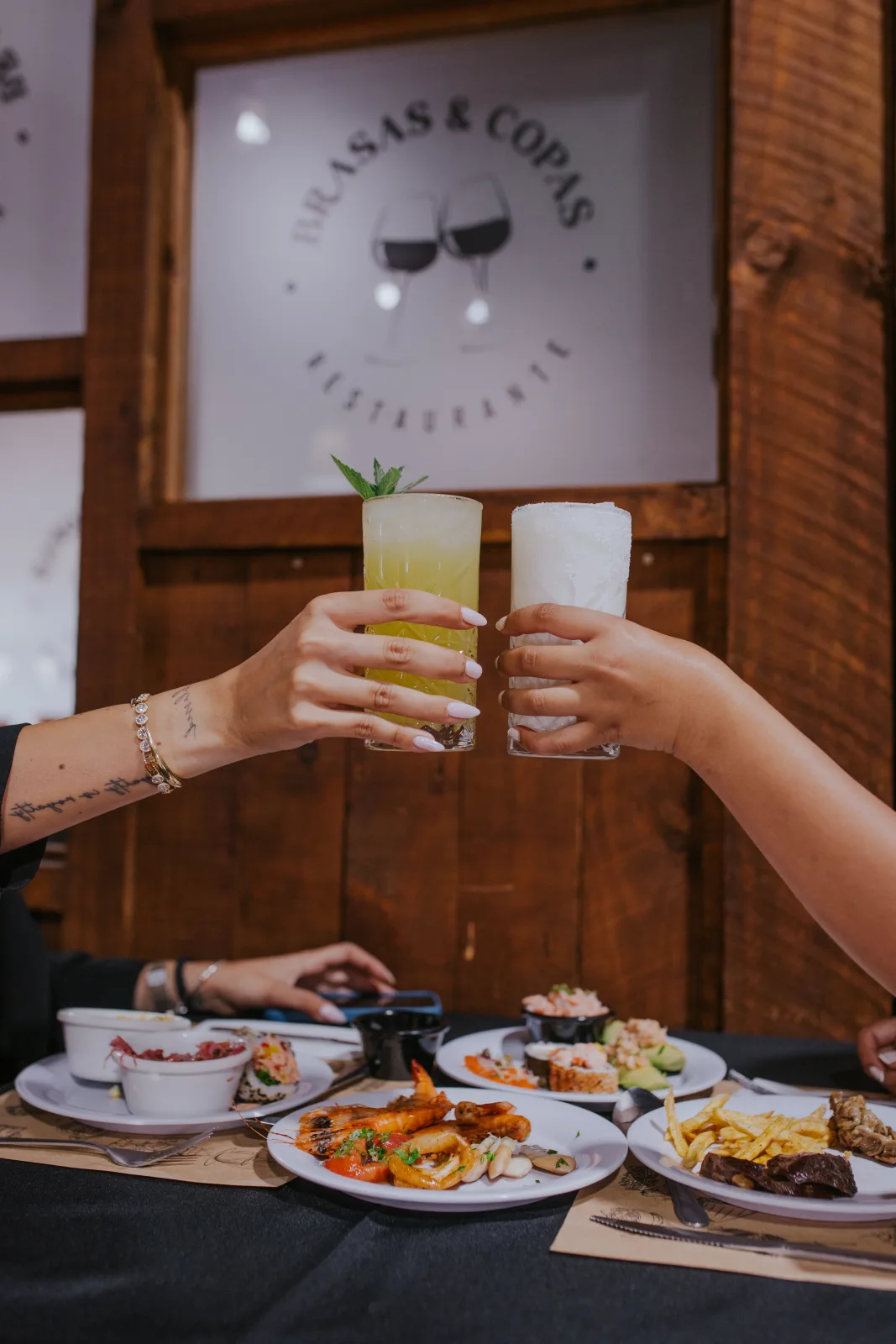 Brindis con tragos sobre una mesa de camarones, ceviche y parrilla bajo el logo de Brasas y Copas
