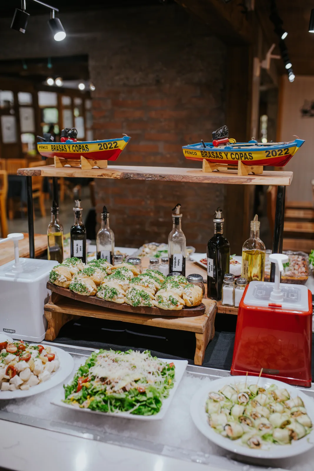 Buffet con panes con perejil, ensaladas frescas y botellas de aceite bajo barquitos decorativos Brasas y Copas