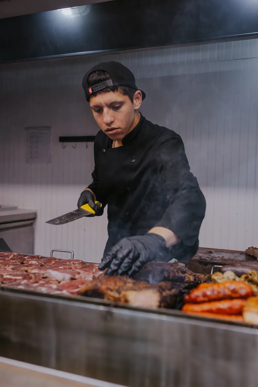 Cocinero parrillero con gorra y guantes negros atendiendo cortes de carne y chorizos en la parrilla