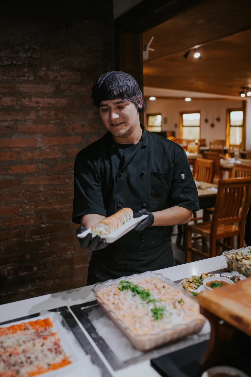 Chef joven con gorro sostiene un roll de sushi flameado sobre la estación fría de mariscos del buffet