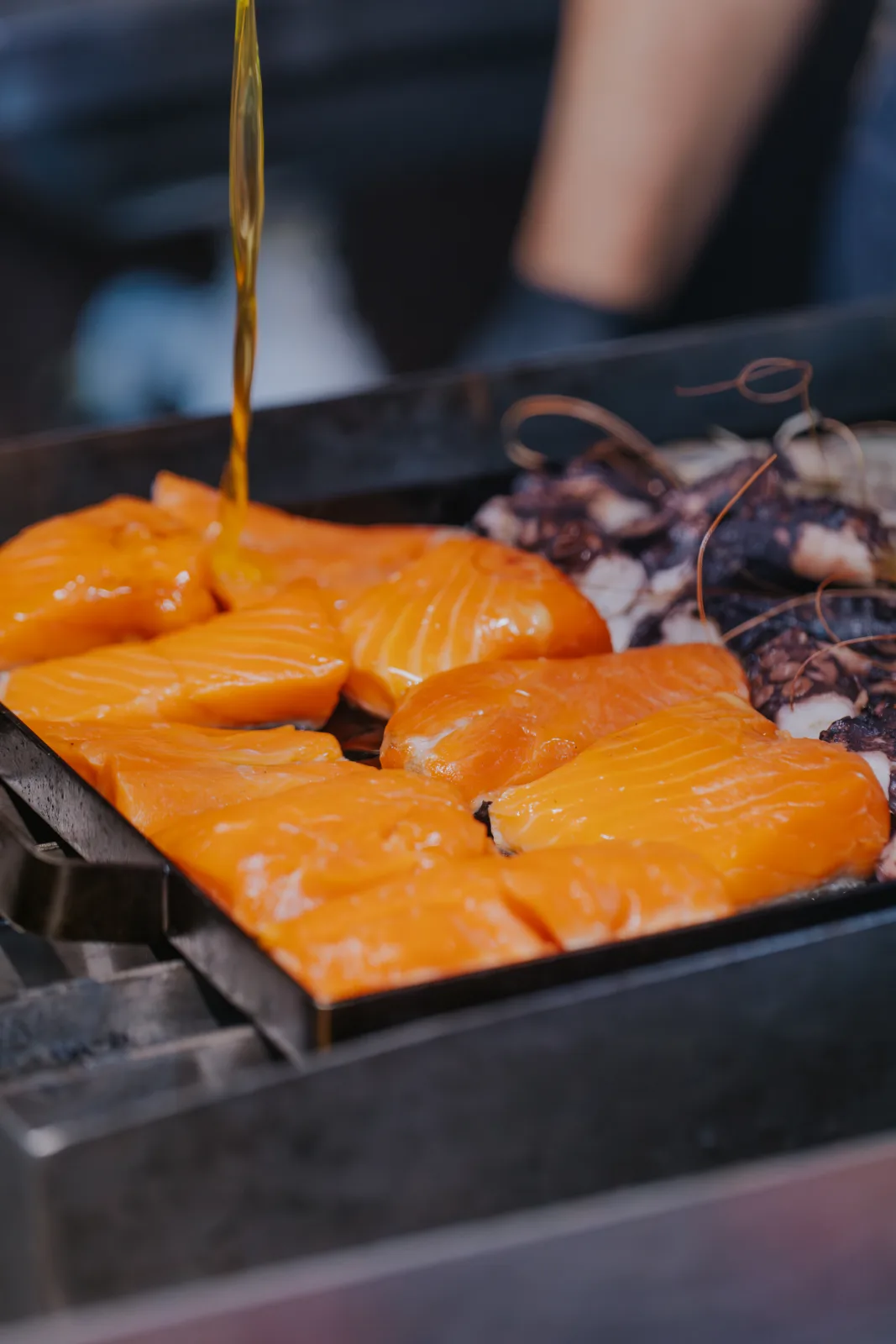 Filetes de salmón fresco en plancha recibiendo un chorro de aceite, con pulpo cocinándose detrás