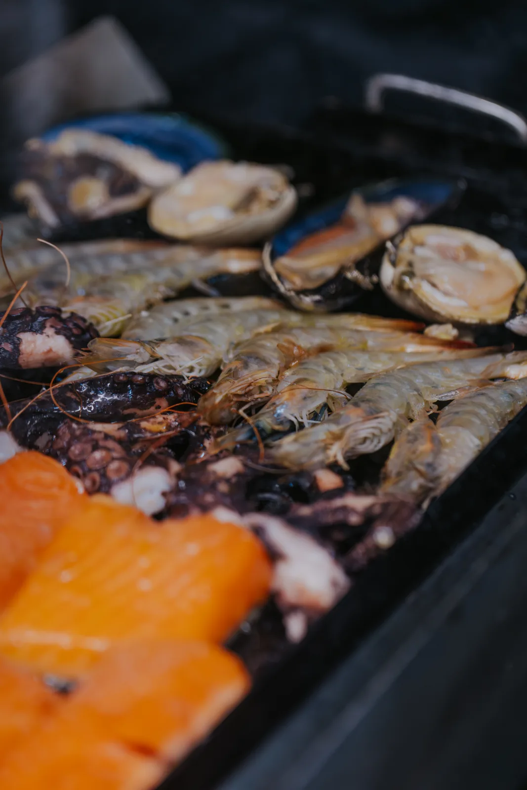 Primer plano de camarones enteros, pulpo y machas frescas asándose junto a salmón en la plancha