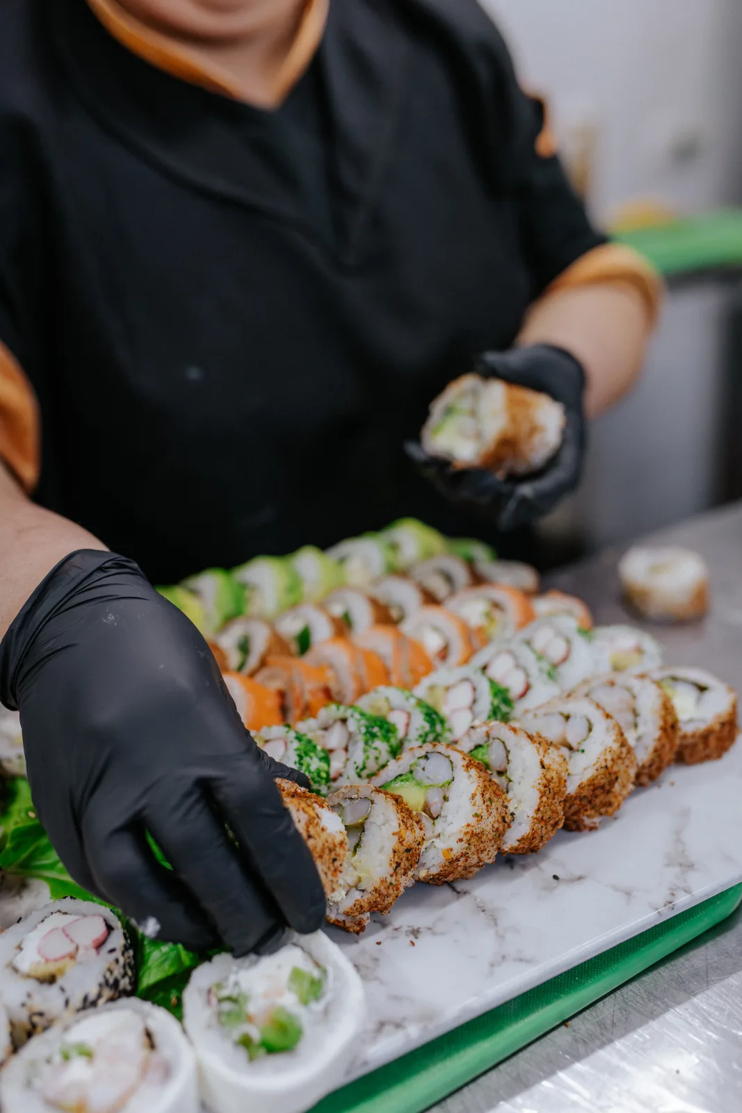 Sushiman con guantes negros ordena hileras de rolls de sushi con salmón y palta sobre tabla de mármol