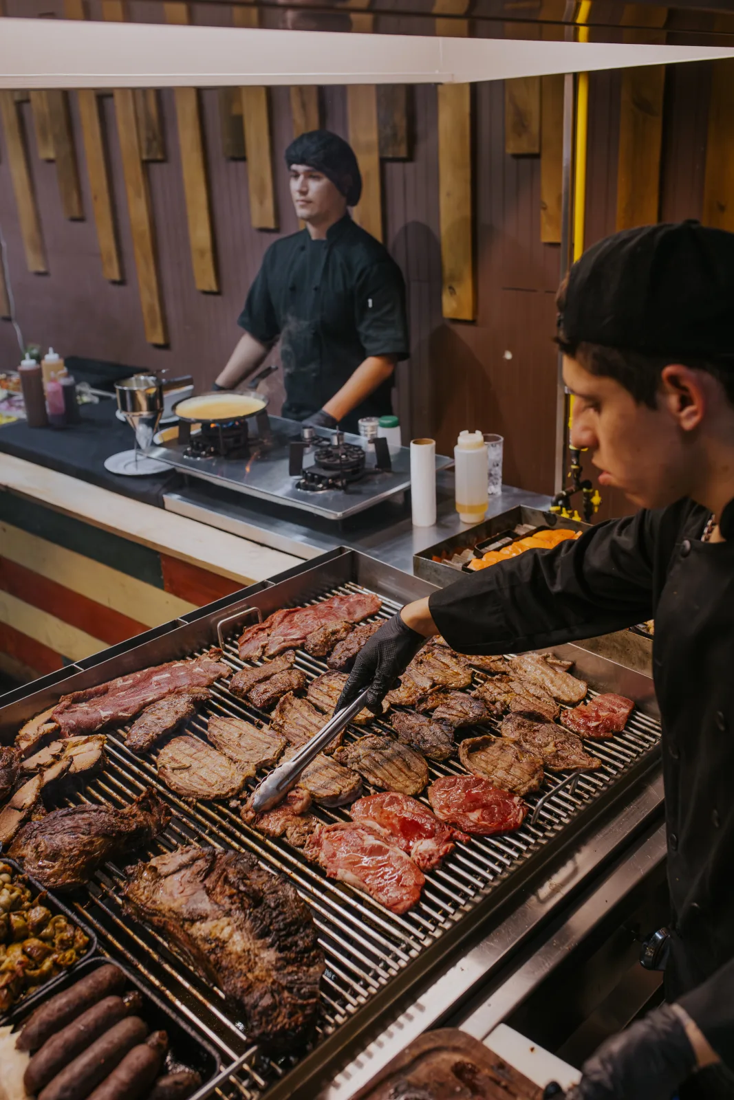 Gran parrilla con cortes de carne y tomates asados atendida por dos cocineros en la estación de brasas